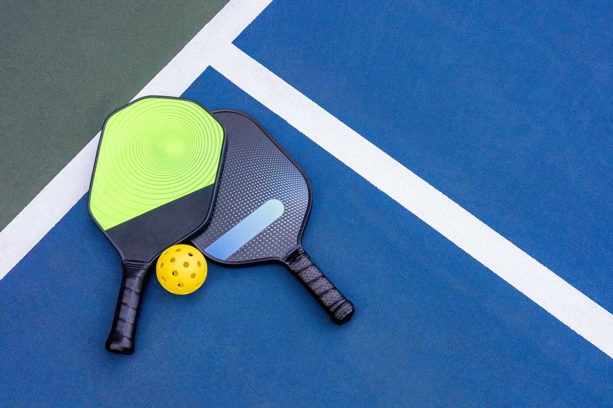 Two pickleball paddles and a ball sit near the sideline of a pickleball court.