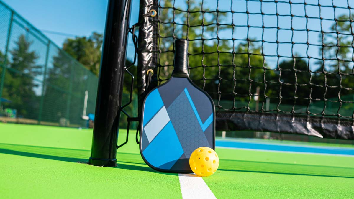 A pickleball paddle and ball rest against the net on a pickleball court. 