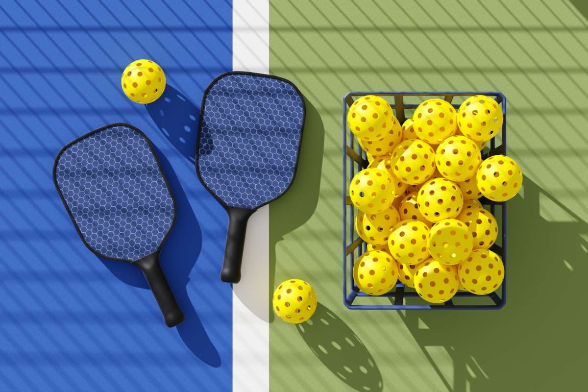 A basket of pickleballs sits next to two rackets on a pickleball court. 
