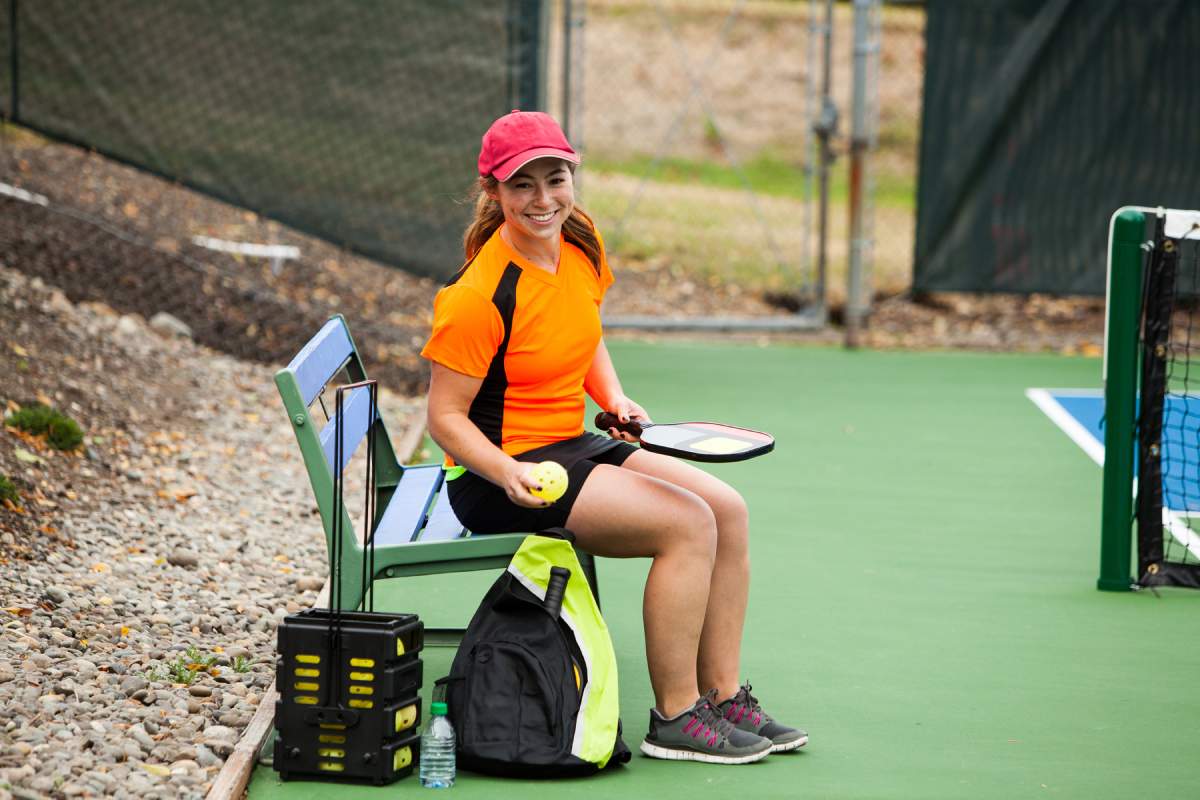 A pickleball player sits on a bench, next to her pickleball bag. 
